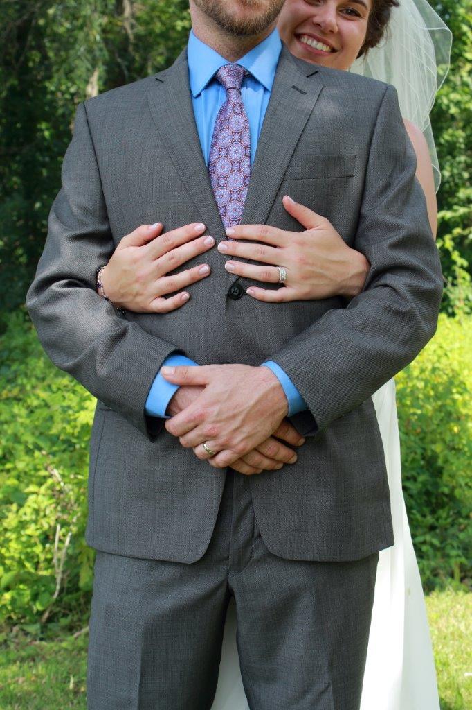 A smiling newly-wed couple holding each other with the bride holding the groom around the waist from behind