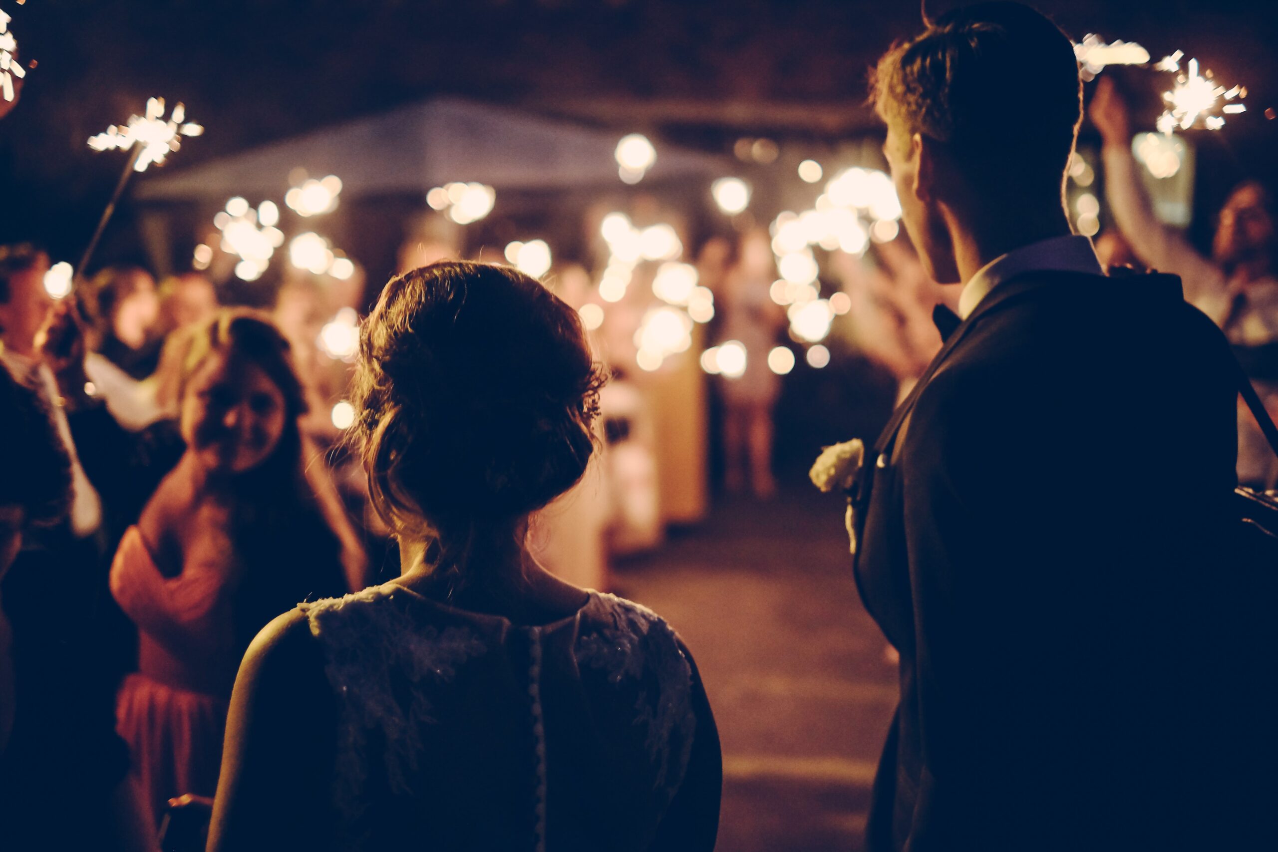 Two people look on at an outdoor nighttime wedding illuminated by sparklers.